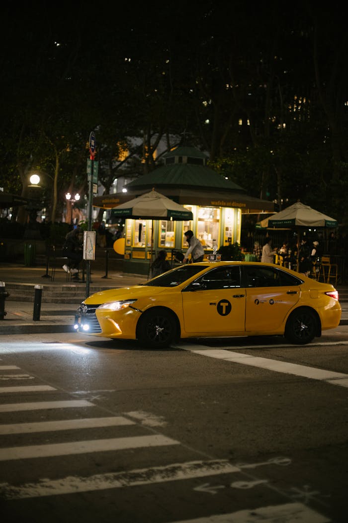 A yellow taxi drives through a dimly lit urban street at night with blurred city lights.
